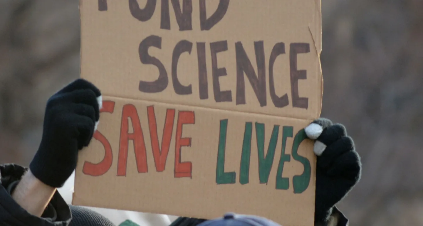 A protest sign reads "FUND SCIENCE SAVE LIVES"