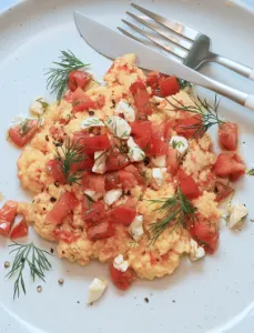 white plate with scrambled eggs, tomatoes and fetta and fork and knife