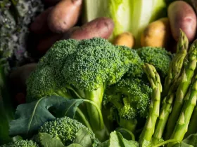Seasonal produce - a closeup of broccoli and asparagus in the foreground, with potatoes, cabbage, collard greens and kale in the background.