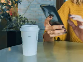 person looking at phone sitting at a cafe table with to-go coffee cup