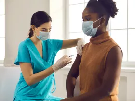 healthcare worker in blue scrubs giving a woman a vaccine shot in her upper arm in front of a bright window