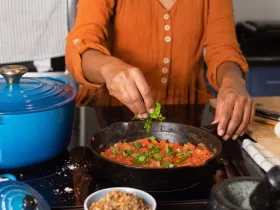 A woman in an orange shirt tears basil over a pot of tomato sauce