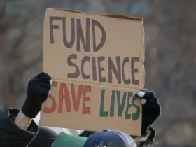 A protest sign reads "FUND SCIENCE SAVE LIVES"