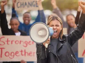 A woman with a raised fist cheers into a microphone; the crowd behind her holds a sign reading "Stronger Together"