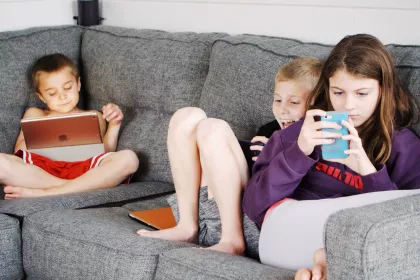 Three young children sit on a couch, looking at phones and tablets