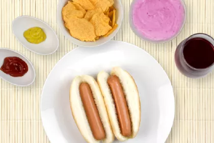 2 hot dogs on a plate surrounded by dips, bowl of chips, blueberry yogurt and cranberry juice
