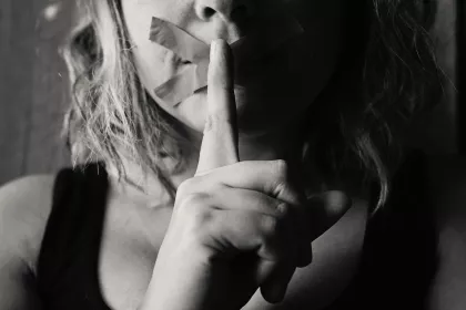 A woman with tape over her mouth making a shush gesture