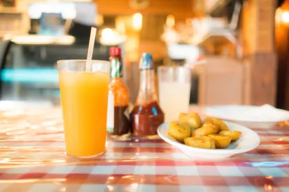 A glass of orange juice with a straw next to a plate of sweet plantains on a checkered tablecloth