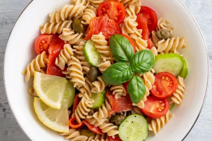 bowl of rotini with cucumber, tomatoes, olives, basil and lemon wedges