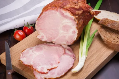 Sliced ham on a wooden cutting board beside two tomatoes, a knife, a scallion, and rustic brown bread.