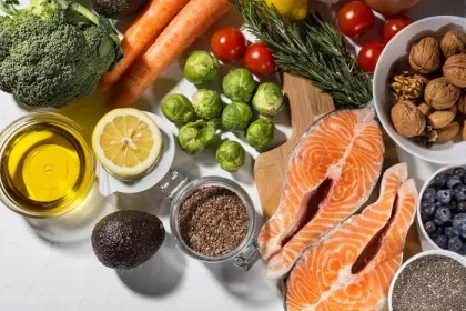 A flatlay of healthy dinner ingredients, including salmon, broccoli, Brussels sprouts, olive oil, nuts and seeds, herbs, and other vegetables.