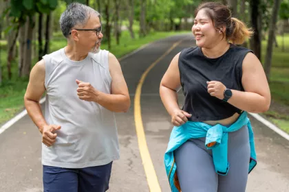 Man and woman with larger bodies unning on a paved trail in a tree lined park