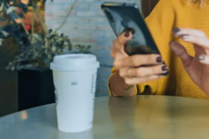 person looking at phone sitting at a cafe table with to-go coffee cup