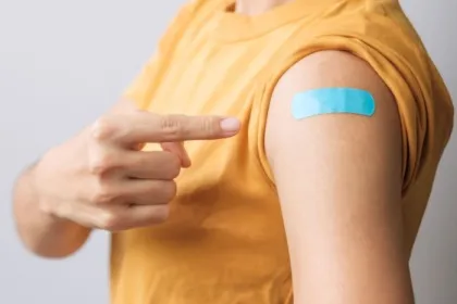 A woman points to her arm, where a blue bandage covers a vaccination injection site