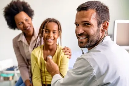 A pediatrician listens to a child's heartbeat with a parent in the background