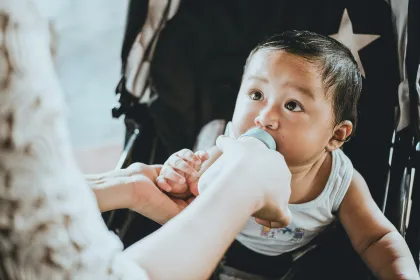 A baby in a stroller being fed from a bottle.