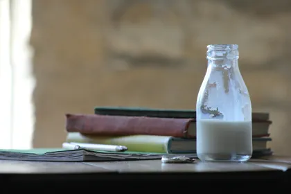 A small bottle of milk on a table beside textbooks and pencils