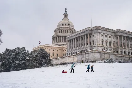 A snowy U.S. Capitol Building with a parent, with their children, pulling a sled uphill 