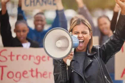 A woman with a raised fist cheers into a microphone; the crowd behind her holds a sign reading "Stronger Together"