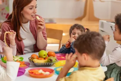 A female teacher sits with young students at a classroom table sharing snacks free from the Big Nine allergens. 