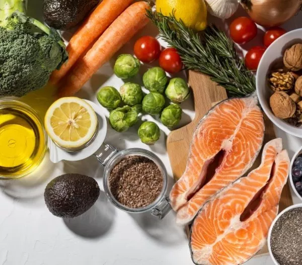 A flatlay of healthy dinner ingredients, including salmon, broccoli, Brussels sprouts, olive oil, nuts and seeds, herbs, and other vegetables.