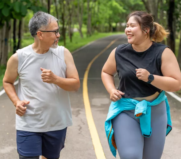 Man and woman with larger bodies unning on a paved trail in a tree lined park