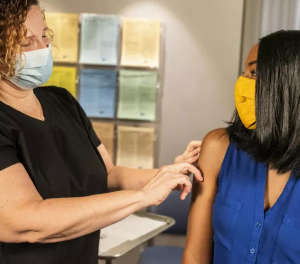 A healthcare worker applying a bandage to a patient after administering a vaccine. Both women are wearing masks.