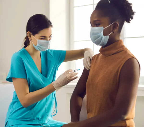 healthcare worker in blue scrubs giving a woman a vaccine shot in her upper arm in front of a bright window
