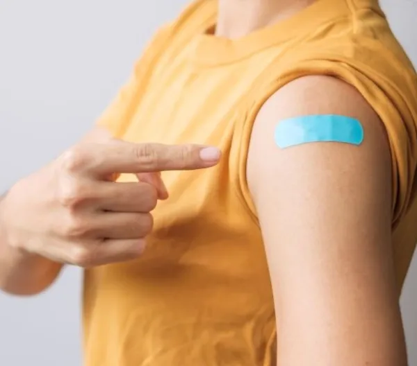 A woman points to her arm, where a blue bandage covers a vaccination injection site