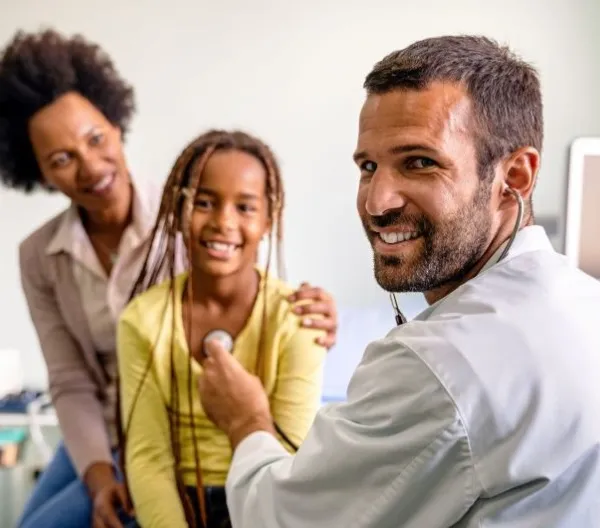 A pediatrician listens to a child's heartbeat with a parent in the background