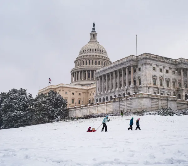 A snowy U.S. Capitol Building with a parent, with their children, pulling a sled uphill 