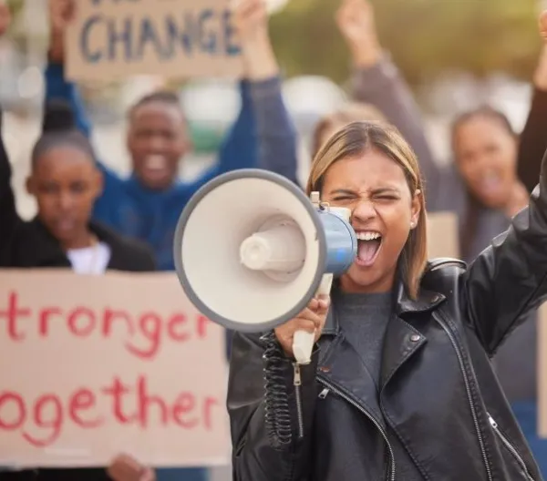 A woman with a raised fist cheers into a microphone; the crowd behind her holds a sign reading "Stronger Together"