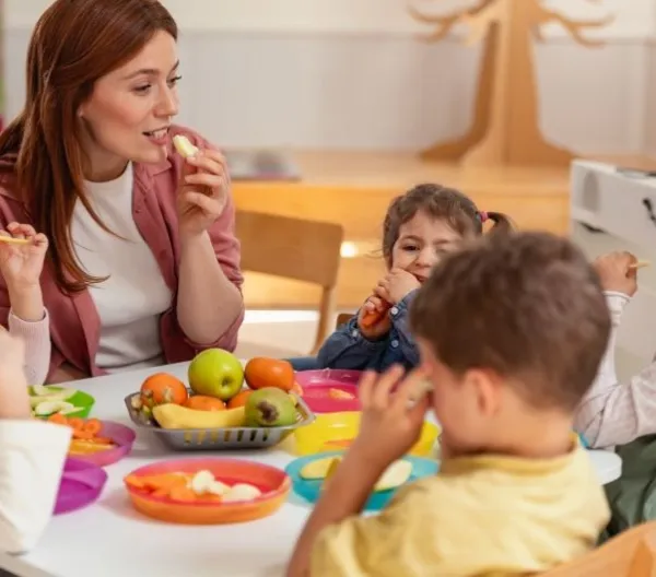 A female teacher sits with young students at a classroom table sharing snacks free from the Big Nine allergens. 