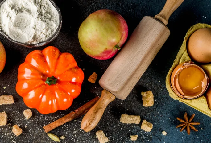 Black background with bowl of flour, small orange pumpkin, apple, rolling pin, carton with one cracked egg, and spices sprinkled in background