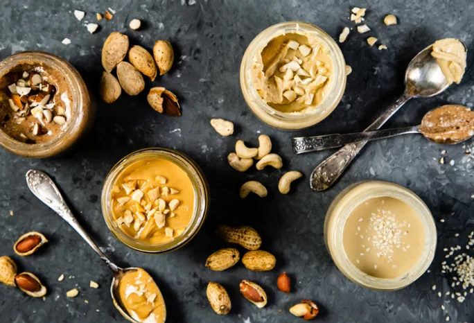 overhead shot of jars of different nut and seed butters with spoons on grey background
