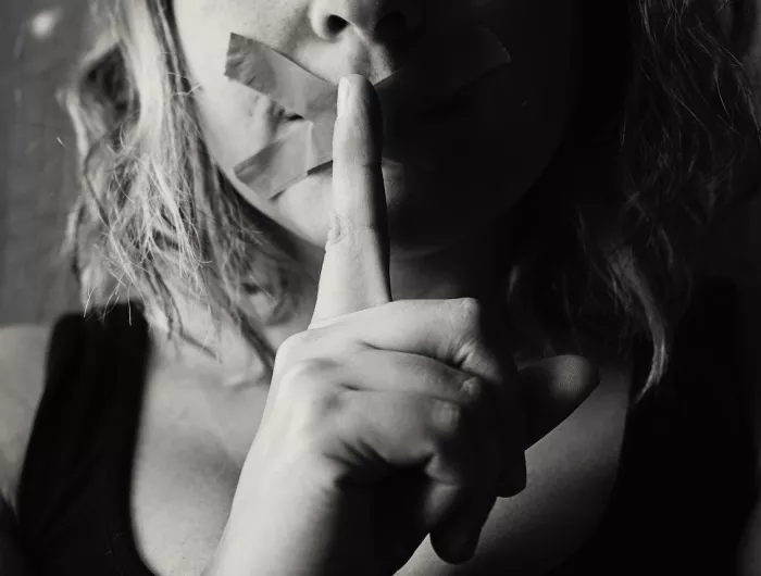 A woman with tape over her mouth making a shush gesture