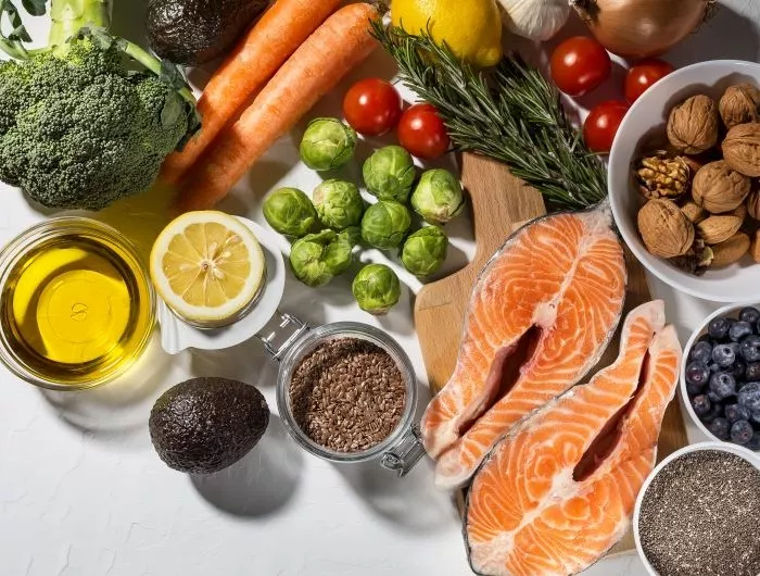 A flatlay of healthy dinner ingredients, including salmon, broccoli, Brussels sprouts, olive oil, nuts and seeds, herbs, and other vegetables.