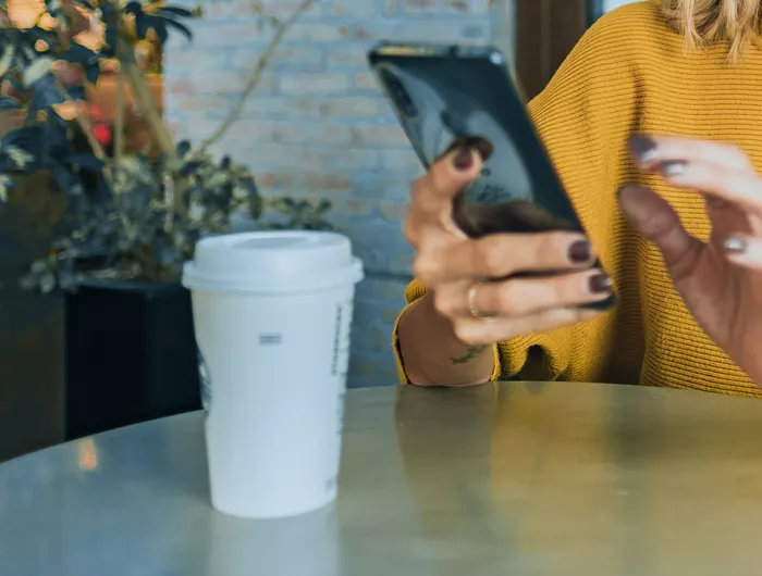 person looking at phone sitting at a cafe table with to-go coffee cup