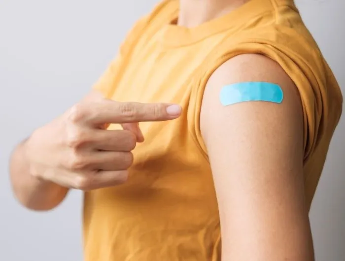 A woman points to her arm, where a blue bandage covers a vaccination injection site