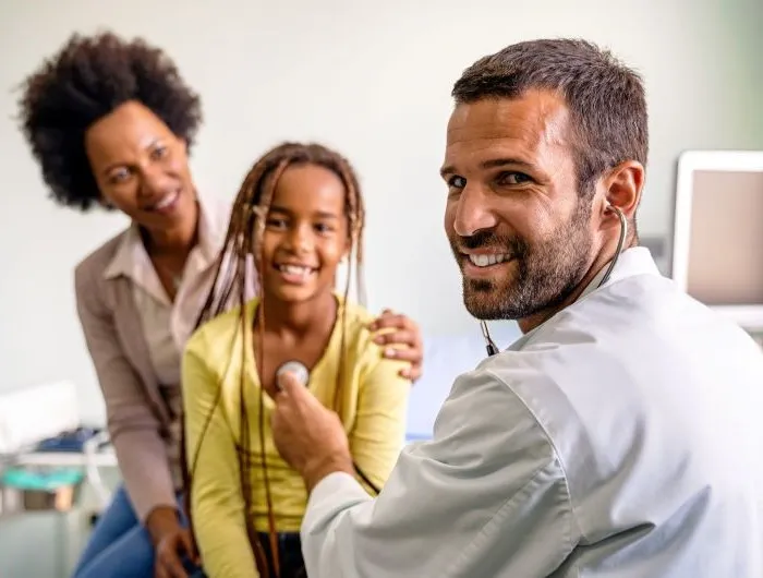 A pediatrician listens to a child's heartbeat with a parent in the background