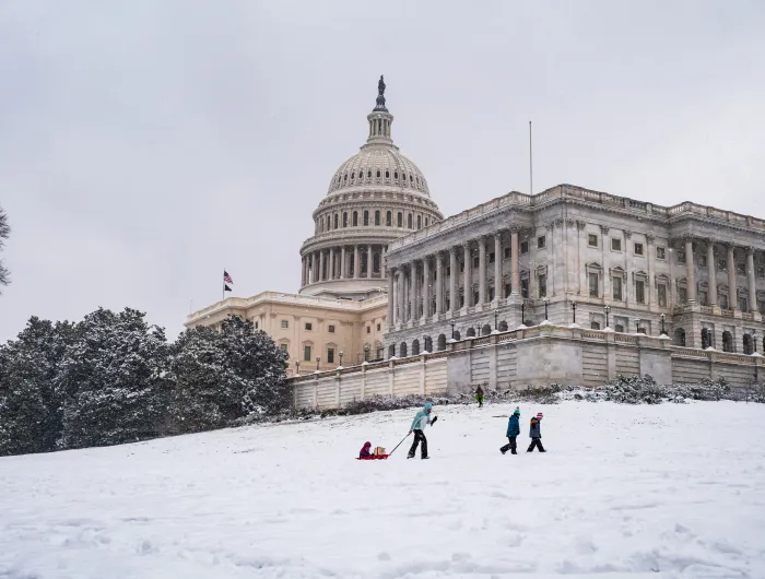 A snowy U.S. Capitol Building with a parent, with their children, pulling a sled uphill 