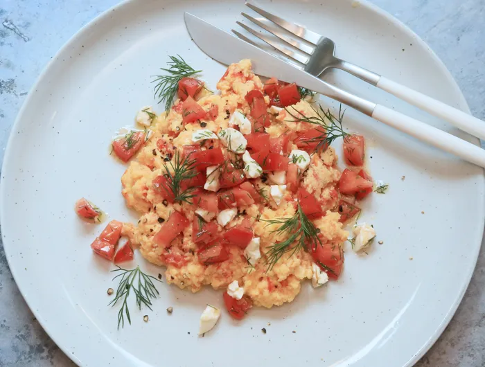 white plate with scrambled eggs, tomatoes and fetta and fork and knife