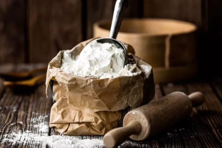 A bag of flour on a wooden table beside a rolling pin