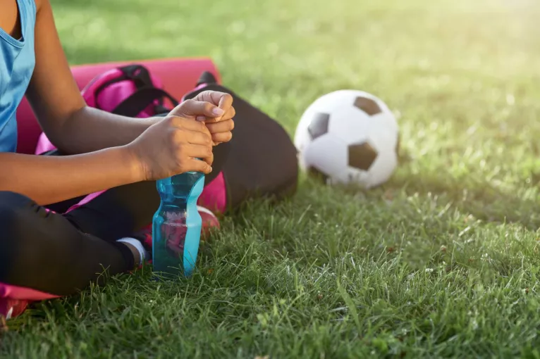 A young Latine girl holds a bottle of water while sitting on a soccer field after practice