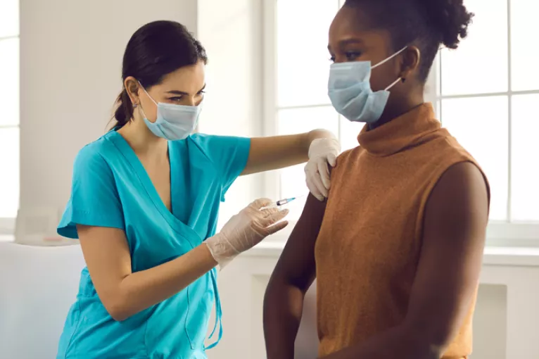 healthcare worker in blue scrubs giving a woman a vaccine shot in her upper arm in front of a bright window