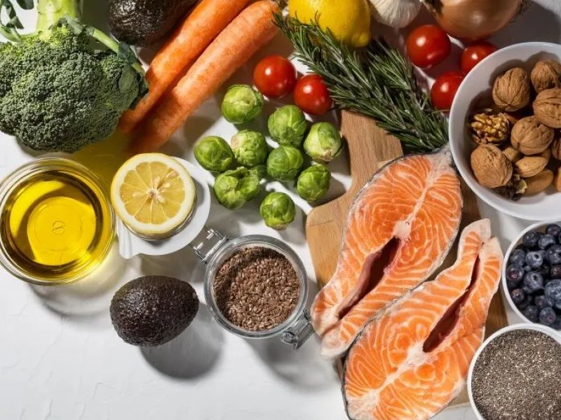 A flatlay of healthy dinner ingredients, including salmon, broccoli, Brussels sprouts, olive oil, nuts and seeds, herbs, and other vegetables.