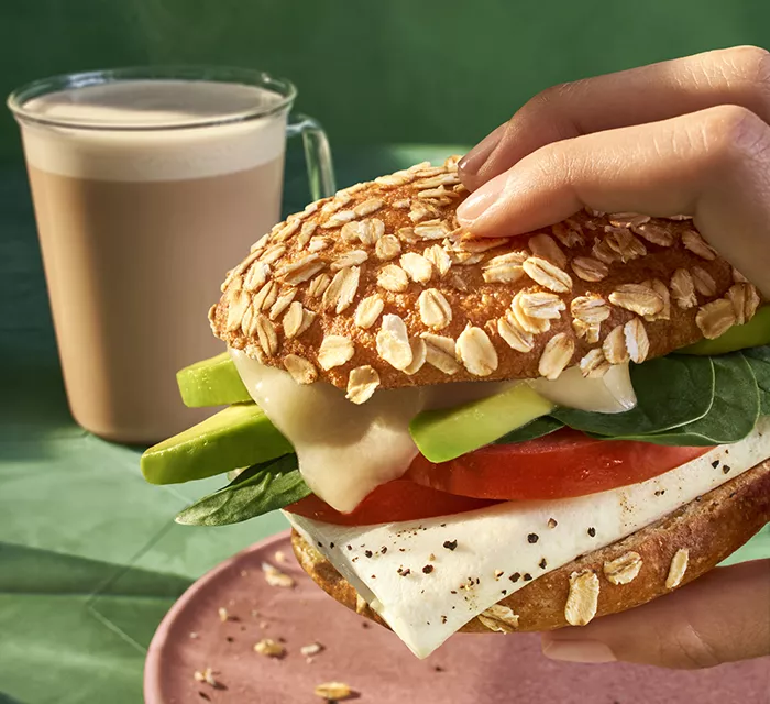 Hand holding Panera's green avo & egg sandwich with coffee in the background