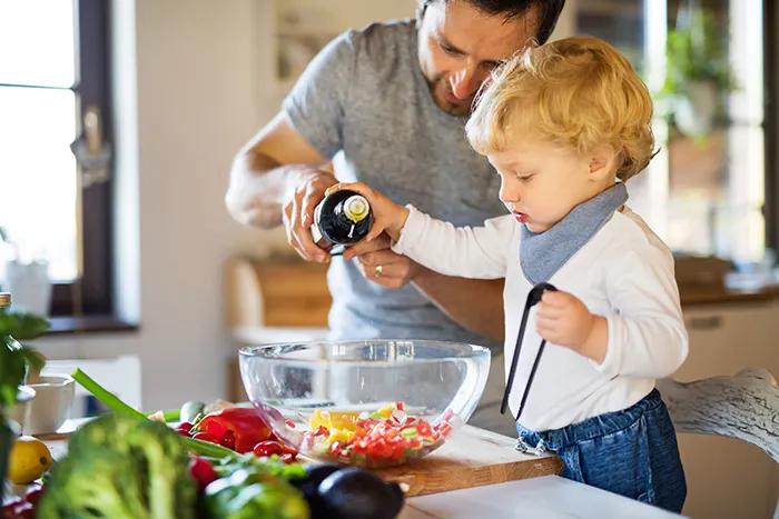 man and toddler in kitchen pouring oil into clear salad bowl with vegetables