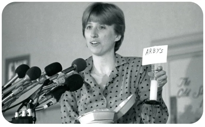 Bonnie speaking into lots of microphones while holding a small sign that says "Arby's"