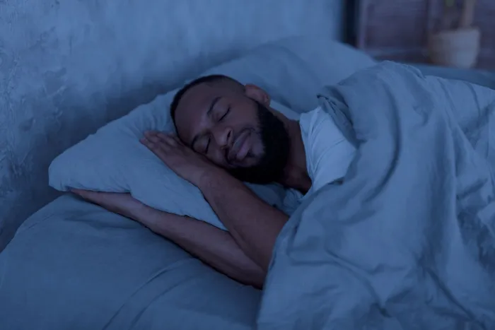 A young Black man sleeps in a dark room and comfortable bed